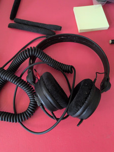 Black headphones on a pink table, things like post it notes are scattered around; the overhead rest is bare and the old cushions lie next to to the headphones